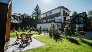 Une maison moderne à deux étages, dotée d’un balcon, entourée d’un jardin bien entretenu avec de petits arbres et d’une terrasse dallée aménagée avec du mobilier d’extérieur.