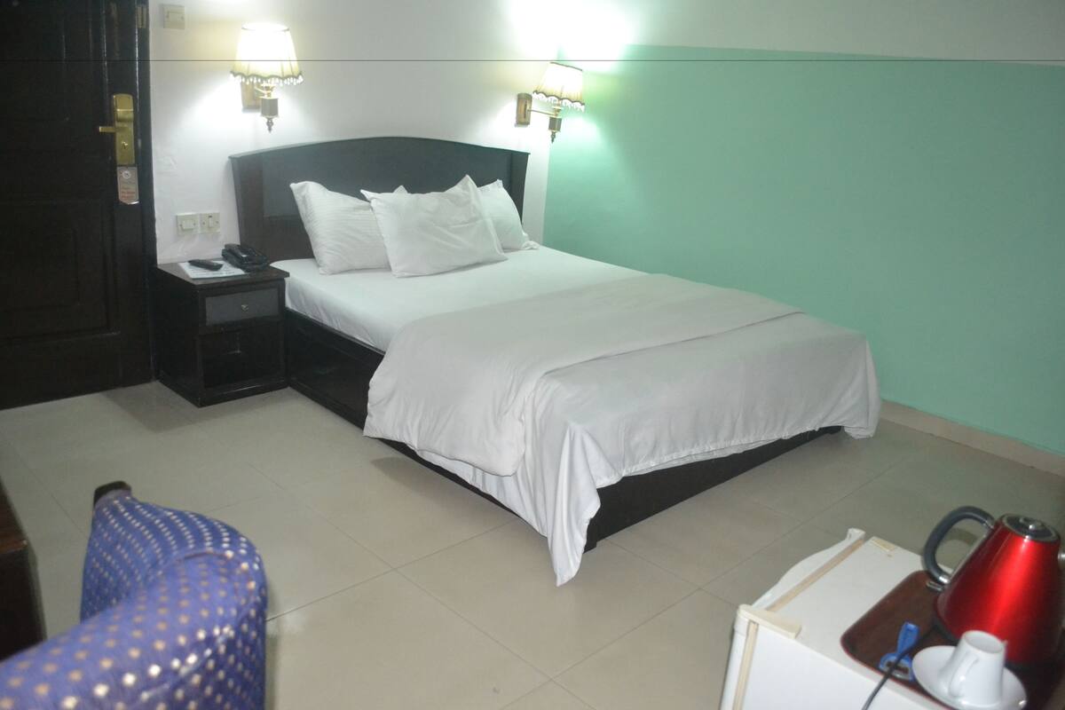 Habitación de hotel con cama, mesita de noche, lámpara y una pequeña mesa con hervidor y taza.