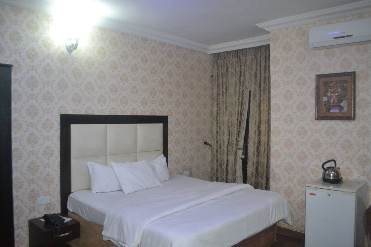 Habitación de hotel con cama, mesita de noche, un pequeño armario y aire acondicionado instalado en la pared.