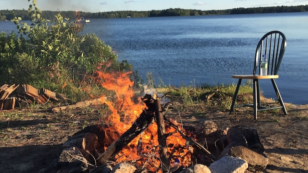 A campfire with logs and rocks, a chair, and a lake with a forested shoreline in the background. - The Shores of Deer Lake (Unorganized Centre Parry Sound)