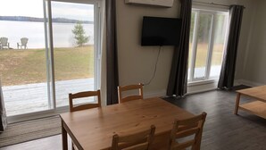 A room with a wooden table and chairs, a flat-screen TV mounted on the wall, and large windows with curtains. - The Shores of Deer Lake (Unorganized Centre Parry Sound)