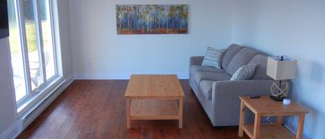 A living room with hardwood flooring, a gray sofa, a wooden coffee table, a side table with a lamp, and a painting on the wall.
