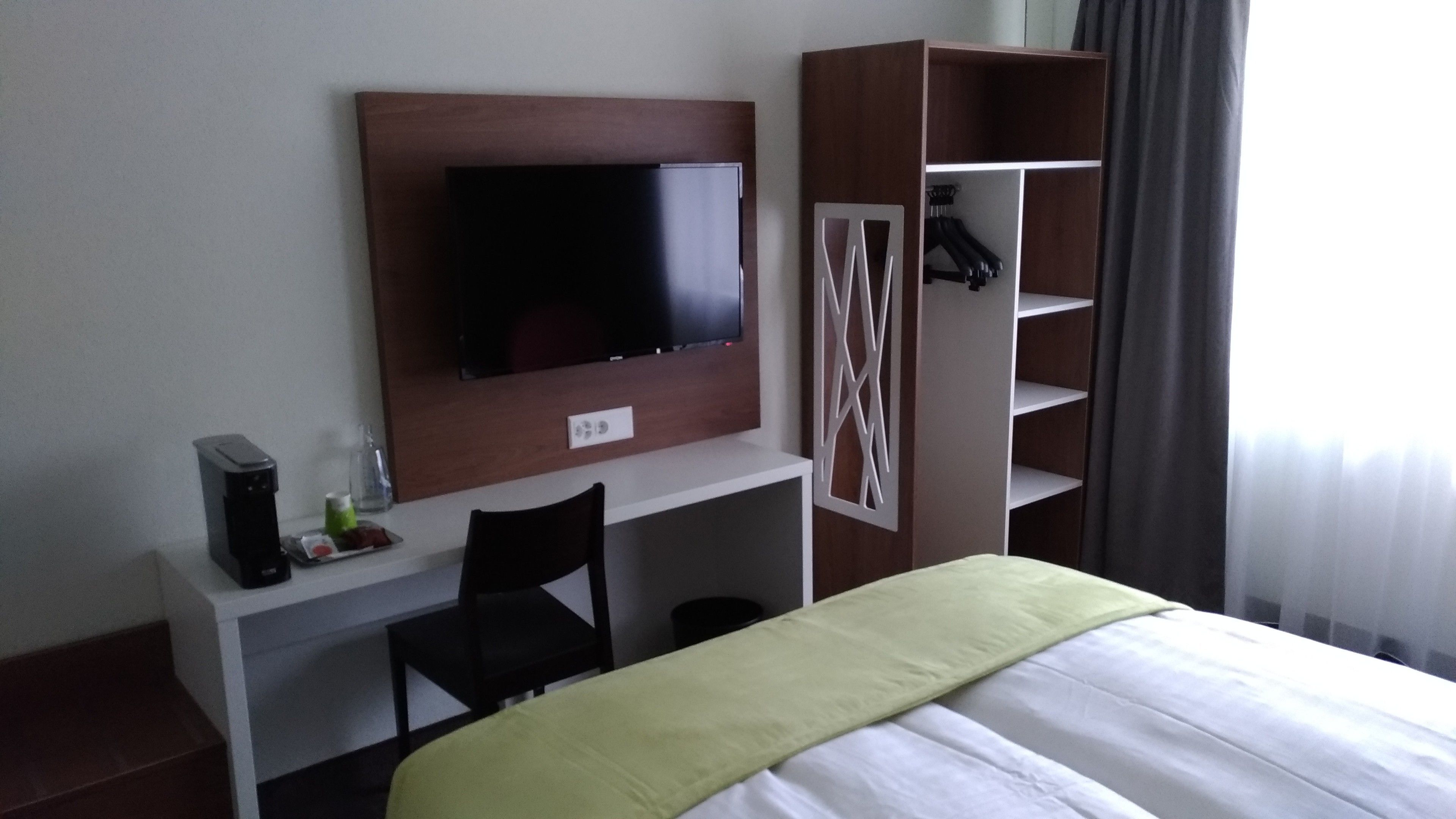 Habitación de hotel con cama, televisor montado en un panel de madera, escritorio con silla y armario con ropa colgada.