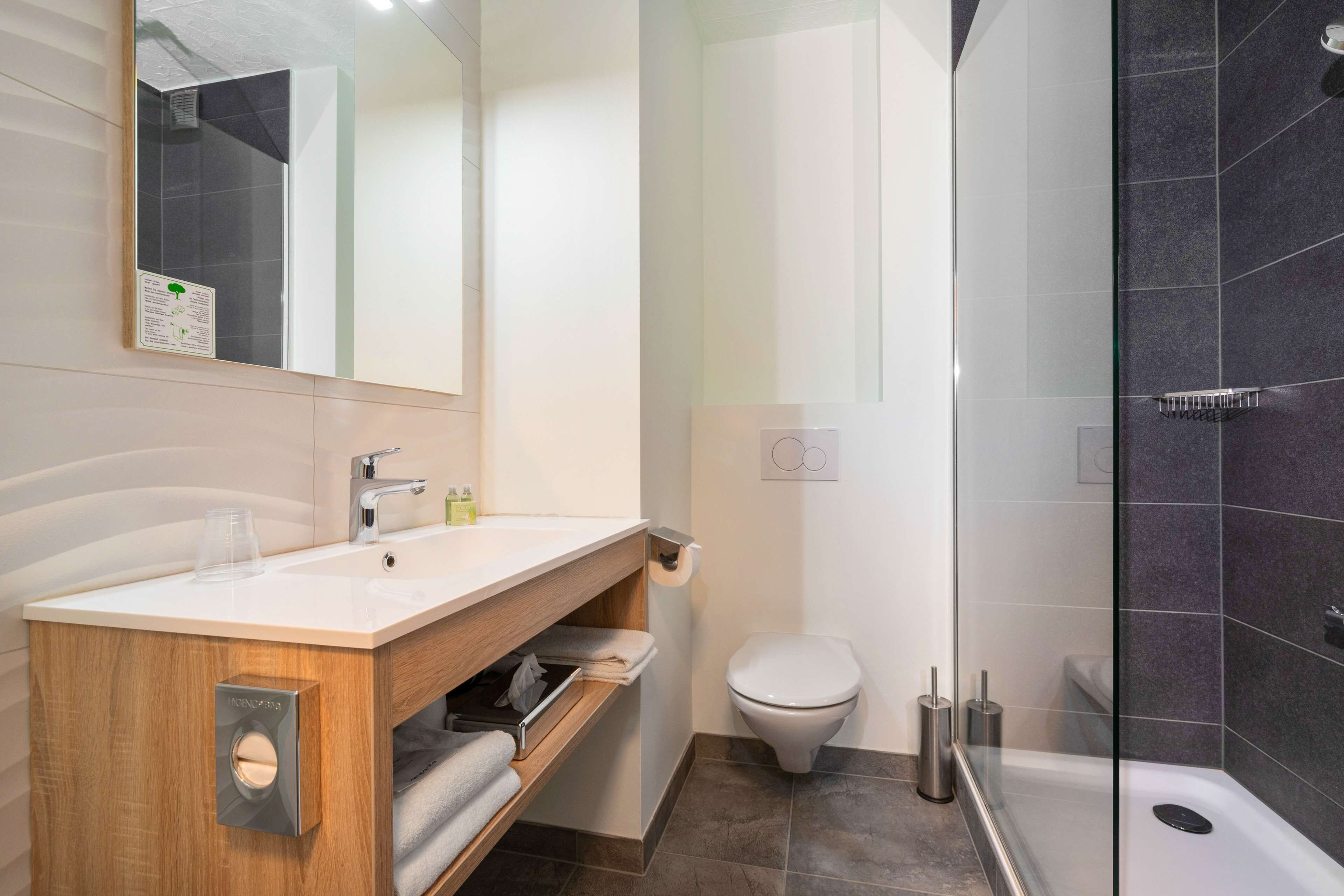 A modern bathroom with a wooden vanity, a white sink, a toilet, and a glass-enclosed shower area.