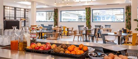 Uma área de café da manhã moderna, com diversas frutas e bebidas no balcão, diferentes opções de assentos e grandes janelas que oferecem vista da cidade.