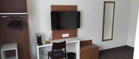 A hotel room with a wooden wardrobe, a desk with a coffee maker, a chair, a TV mounted on the wall, and a mirror.