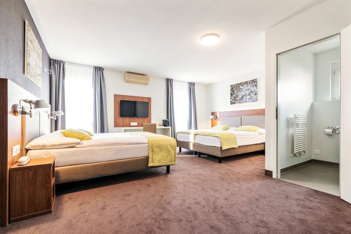 A hotel room with two beds, a TV, a desk, and a bathroom with a radiator.