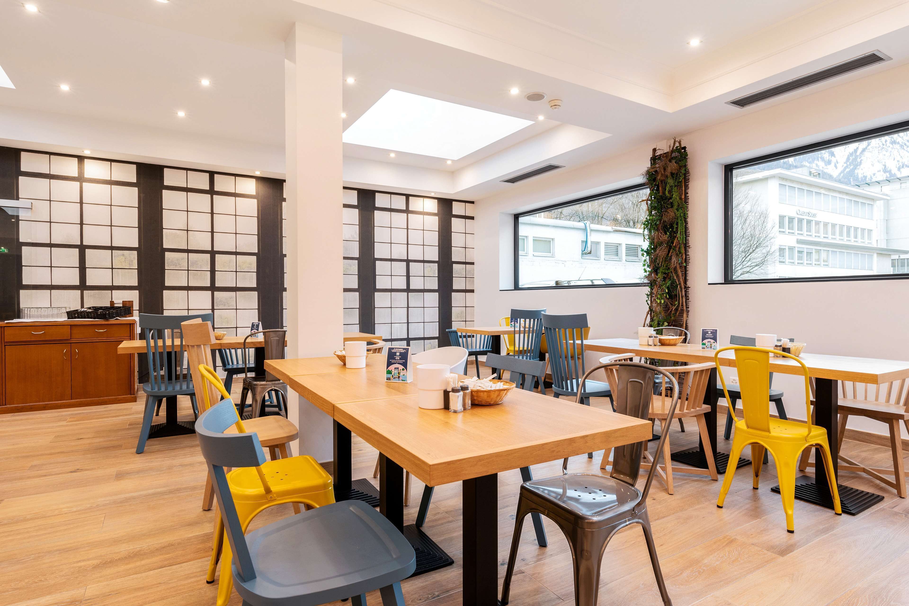 A spacious dining area with multiple tables and chairs, large windows with a view of buildings, and a skylight.