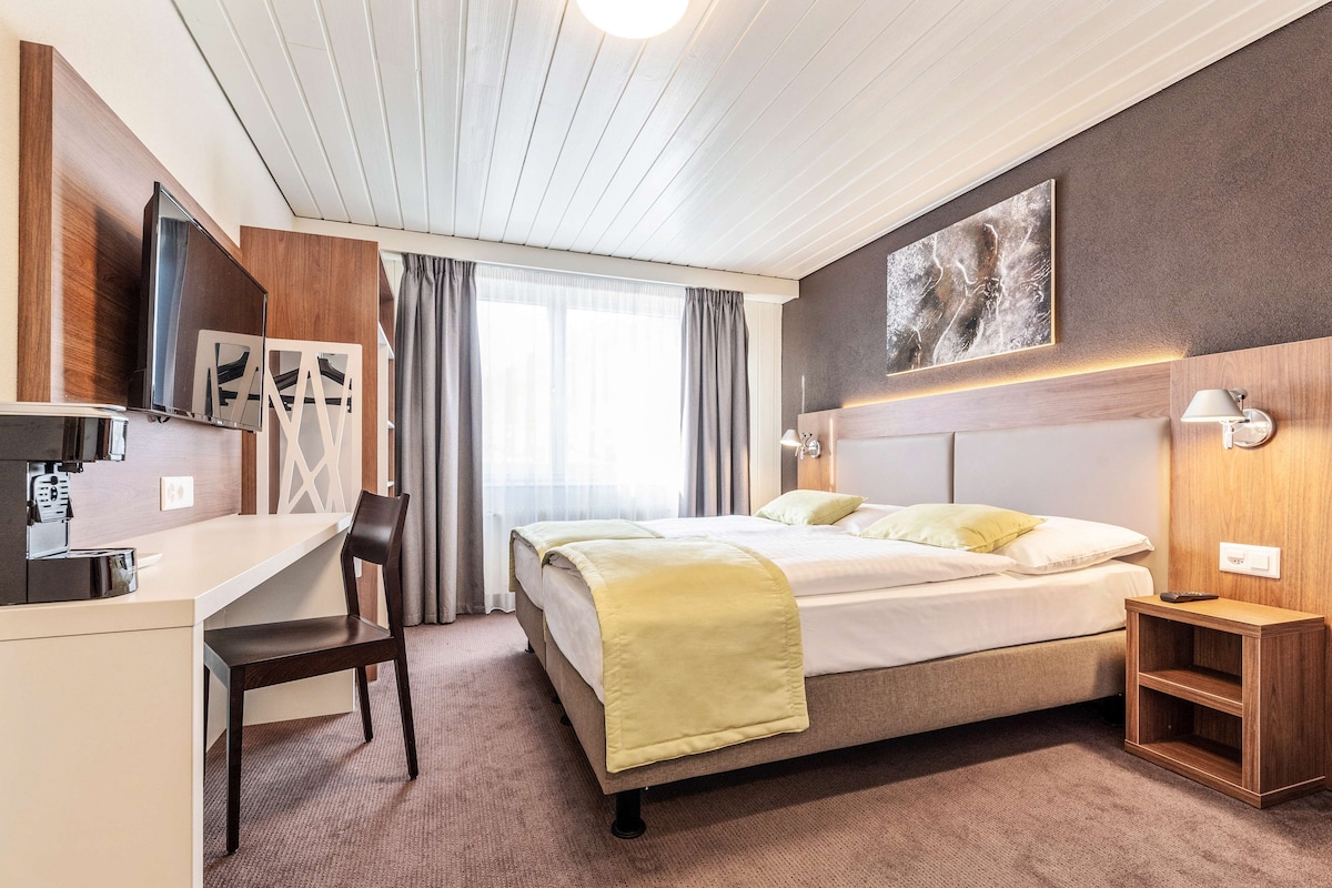 A hotel room with a bed, a desk, a chair, a TV, and a painting on the wall.