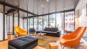 A modern living room with a sectional sofa, two armchairs, a coffee table, and a wooden ottoman. Large windows provide a view of the street. - Campanile Martigny (Martigny)
