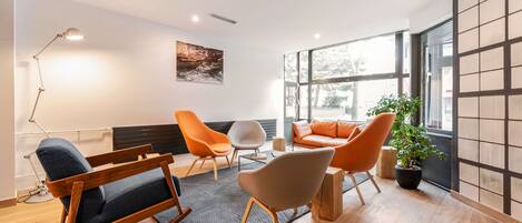 Un salon moderne avec un fauteuil à bascule, des fauteuils orange, un fauteuil gris, une lampe sur pied et une grande fenêtre donnant sur des arbres.
