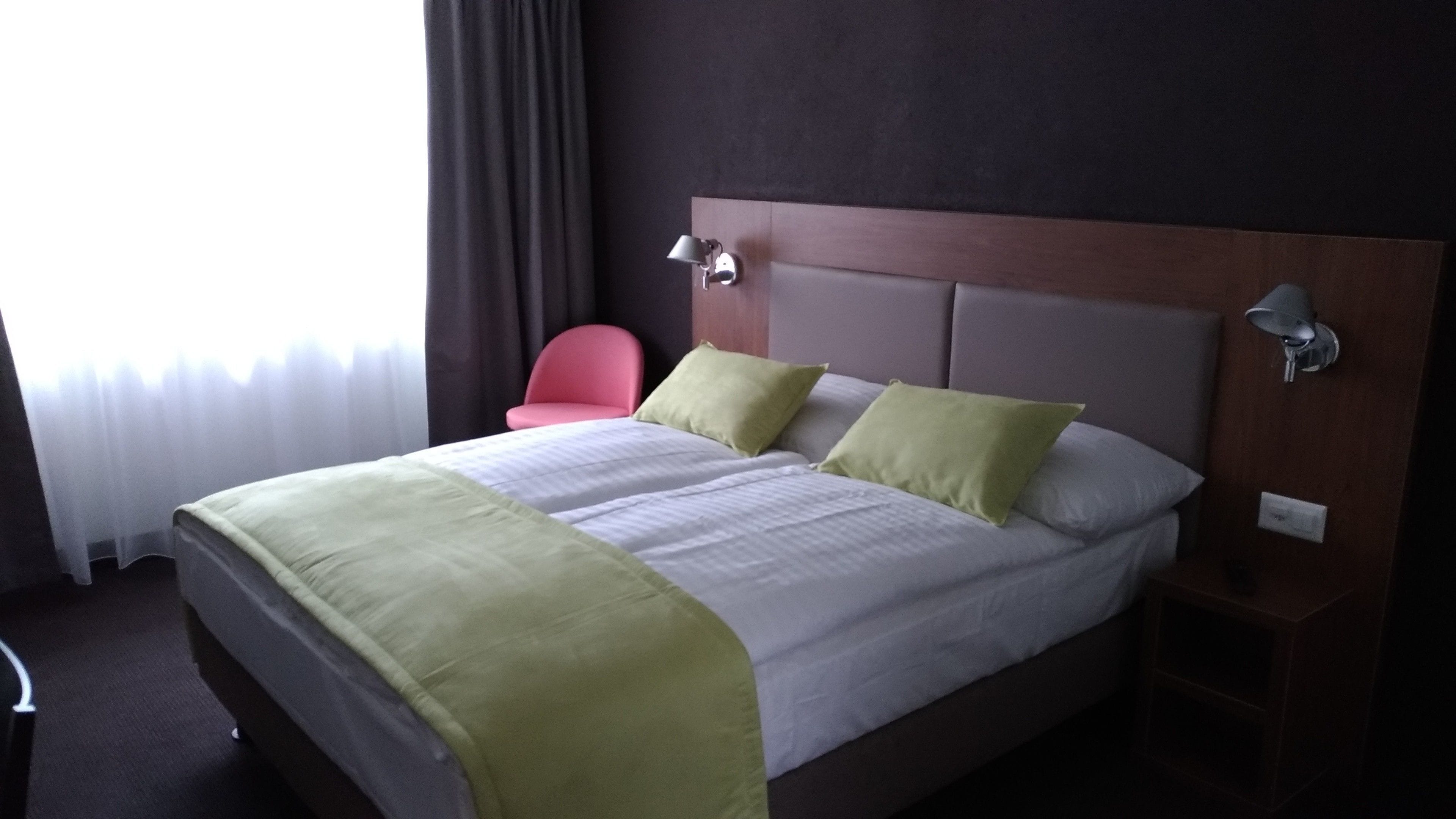 Una habitación de hotel con una cama grande, una mesita de noche, una lámpara, una silla y una ventana con cortinas.
