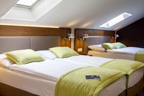 Hotellihuoneessa on kolme sänkyä, ja jokaisessa on vihreä tyyny ja valkoinen peitto. Sänkyjen yläpuolella on kattovalokuvioita, ja huoneessa on puiset päätylevyt sängyissä.
