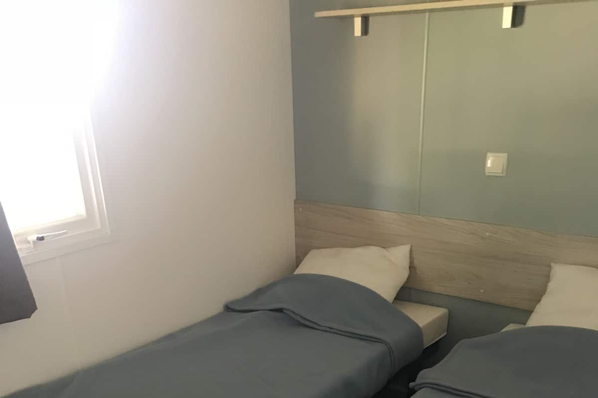 A room with two single beds, a wooden headboard, a window with a curtain, and a light switch on the wall.