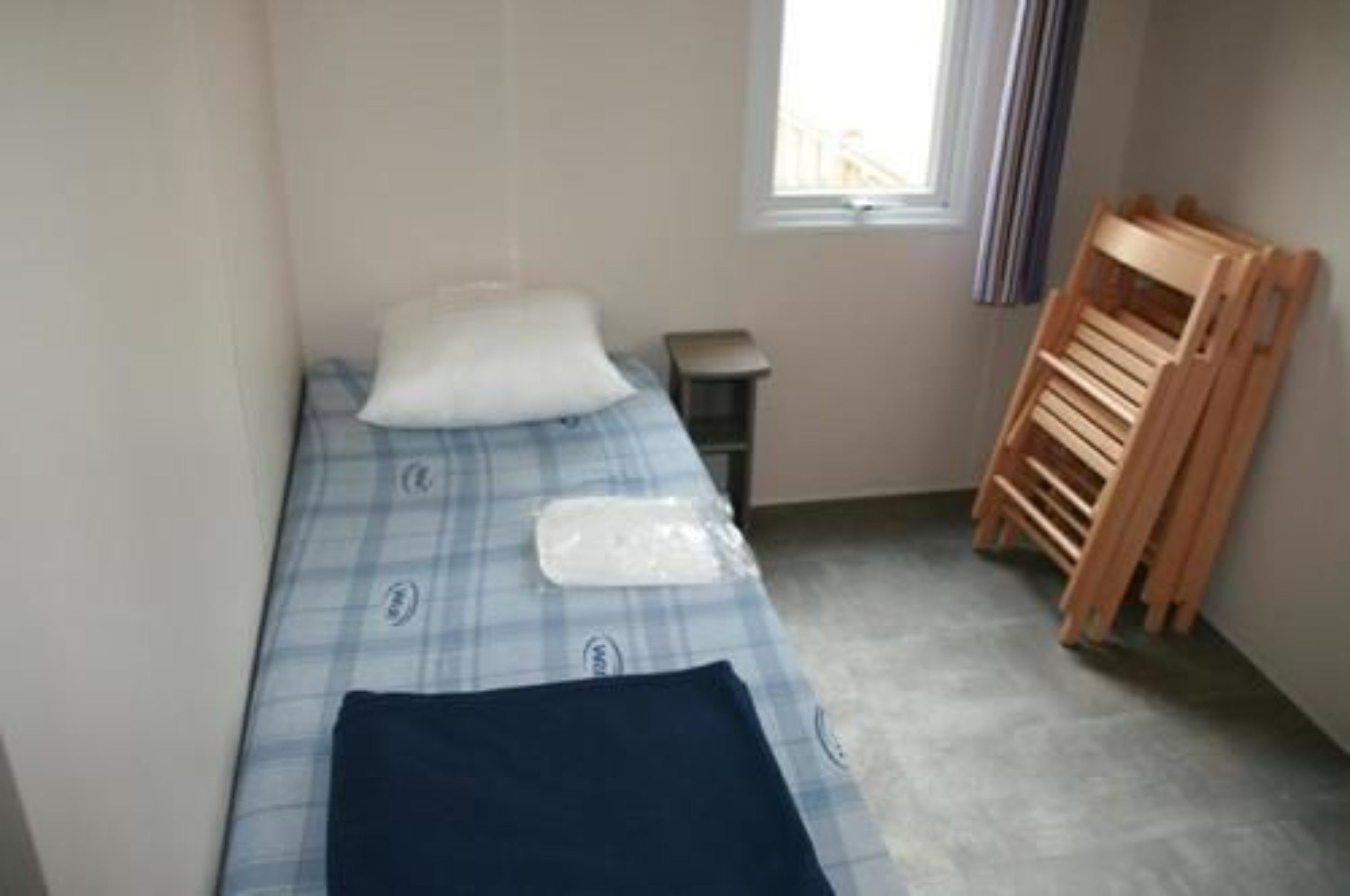 A small, single-bed room with a checkered bedspread, a pillow, a bedside table, and a wooden chair.