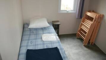 A small, single-bed room with a checkered bedspread, a pillow, a bedside table, and a wooden chair.