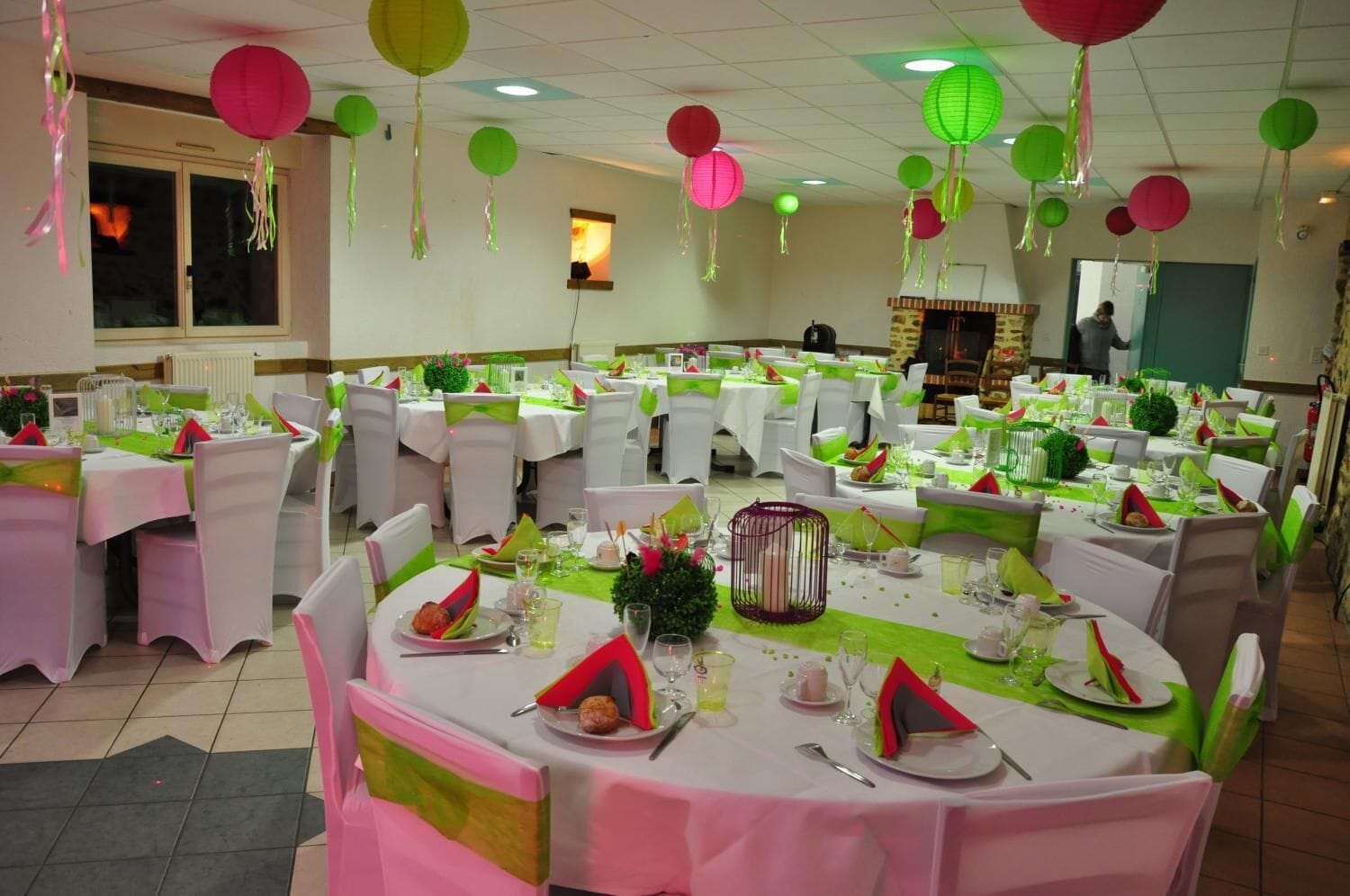 A banquet hall with round tables set for a formal event, featuring green and white tablecloths, white chairs, and hanging paper lanterns.