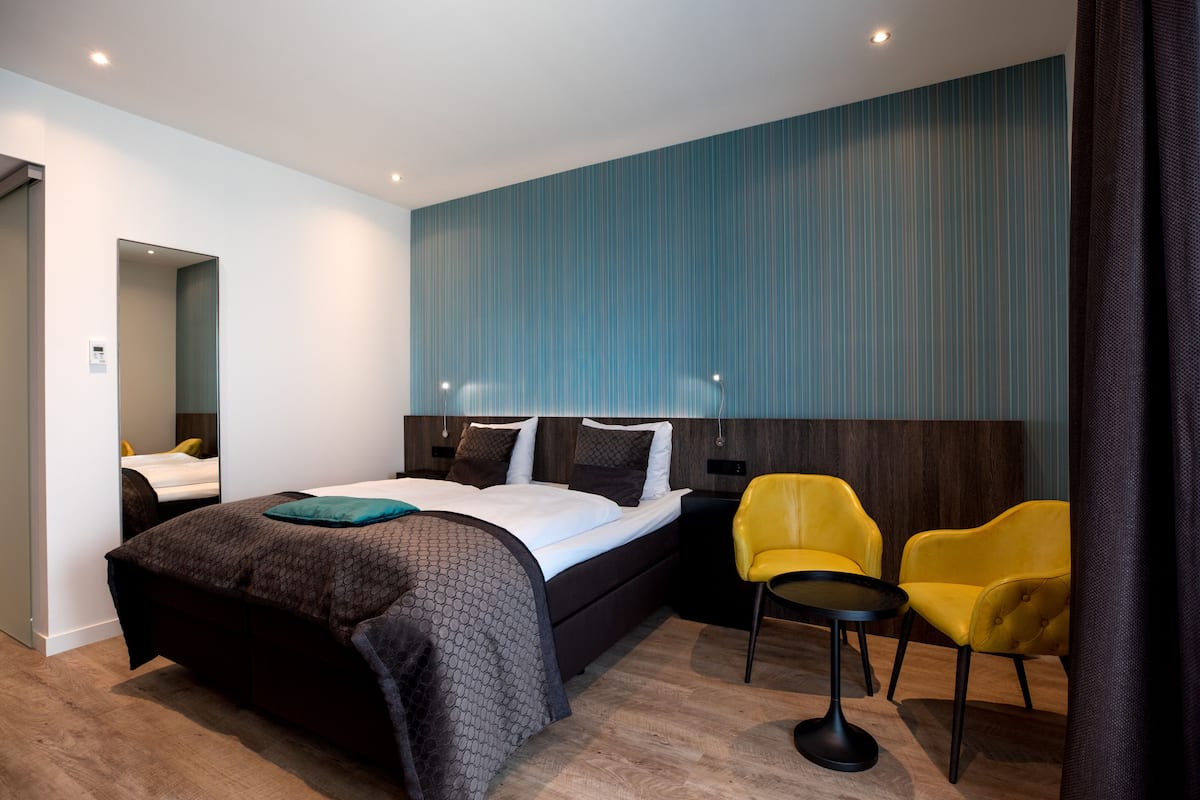 A modern hotel room with a large bed, two yellow chairs, a small table, and a blue-striped wall.