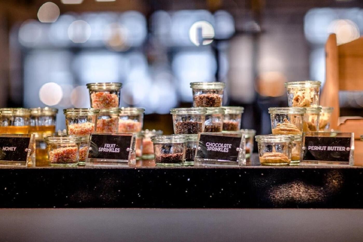 A display of various food items in glass jars labeled with names like honey, fruit sprinkles, chocolate sprinkles, and peanut butter.
