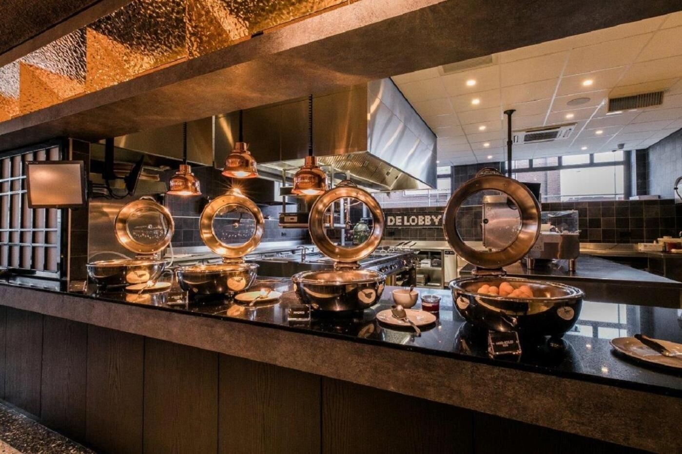 Uma área de buffet com vários recipientes redondos cor de cobre e uma placa visível com a inscrição "DE LOBBY".