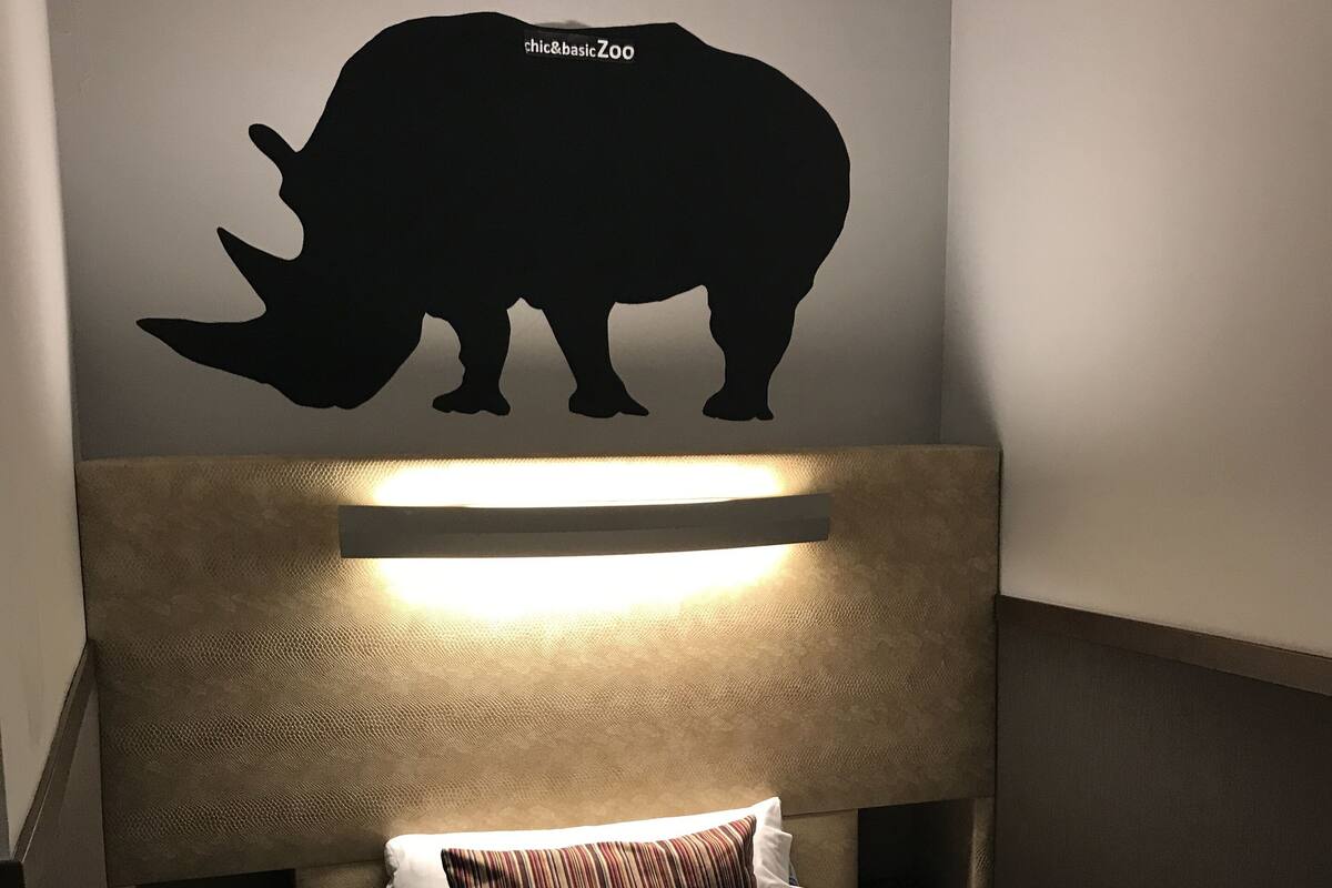 Habitación de hotel con cabecera que presenta la silueta de un rinoceronte, cama con ropa blanca y almohada a rayas.