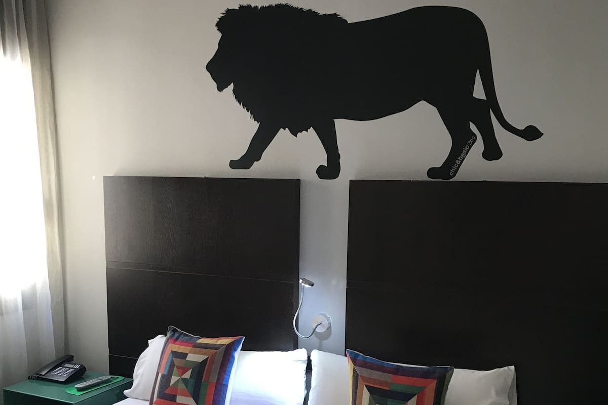 Habitación de hotel con una cama grande, dos almohadas, una mesita de noche con un teléfono y una silueta de león en la pared.