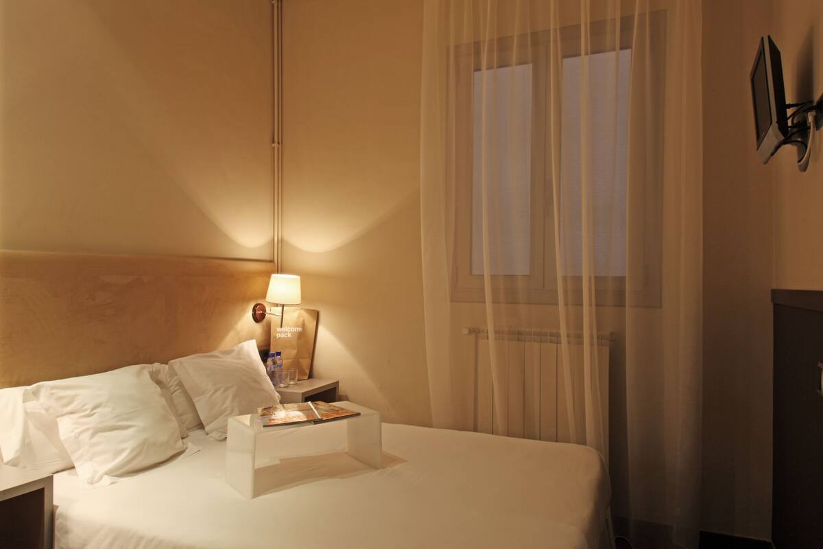 Una habitación de hotel con una cama bien hecha, una lámpara de noche, un mesita de noche y una ventana con cortinas translúcidas.