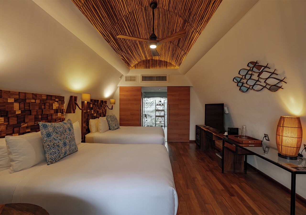 Habitación de hotel con dos camas, ventilador de techo, televisor, mesa con lámpara y panel de pared de madera.