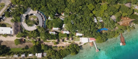 An aerial view of a resort with a circular building, multiple bungalow-style accommodations, and a dock extending into clear turquoise waters.
