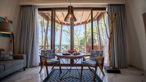 A room with a round table, two chairs, a sofa, and a view of palm trees and the sea.