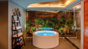 A spa area with two hot tubs, a wooden shelf with towels, and a glass wall with plants.