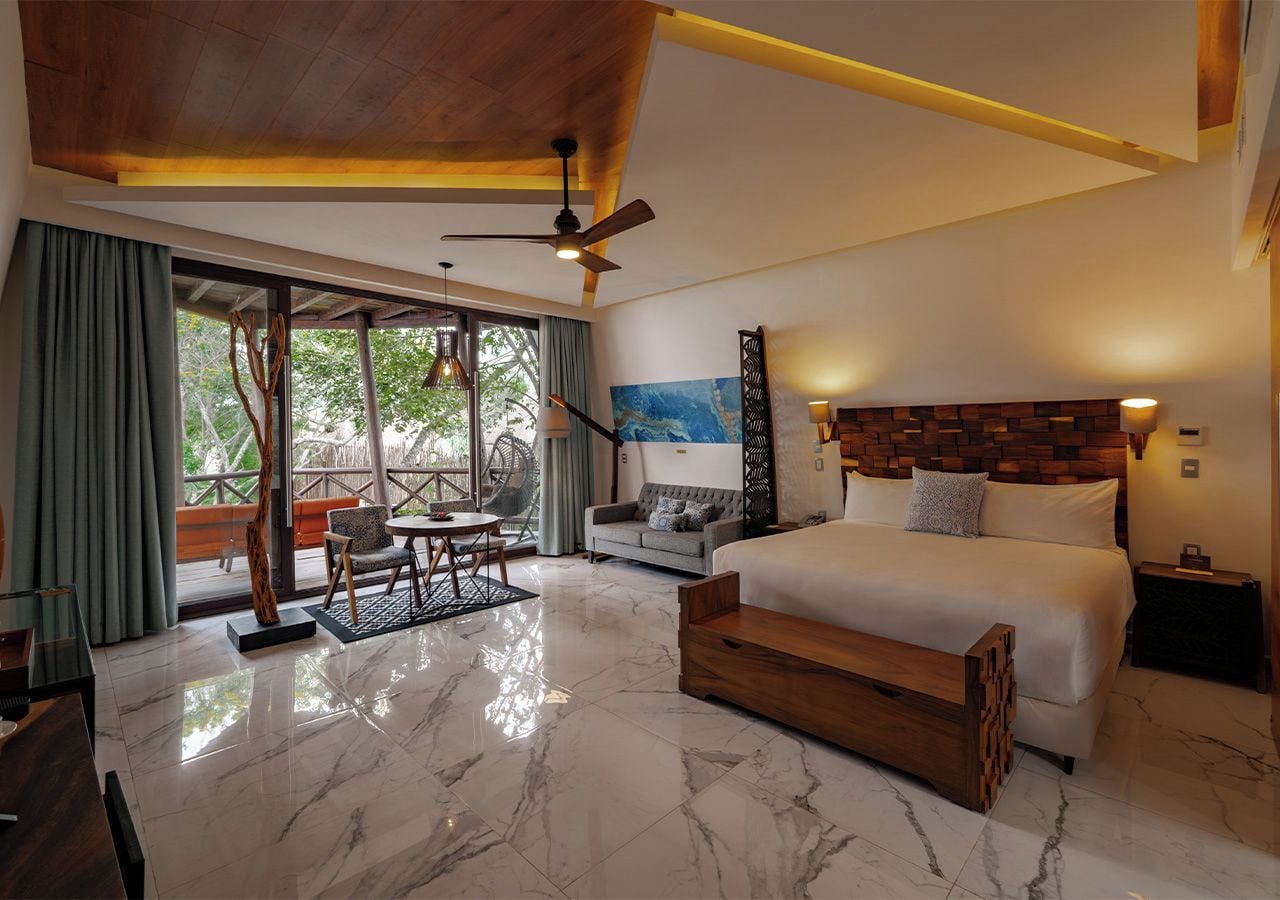 A modern hotel room with a large bed, a seating area, and a view through a glass door.