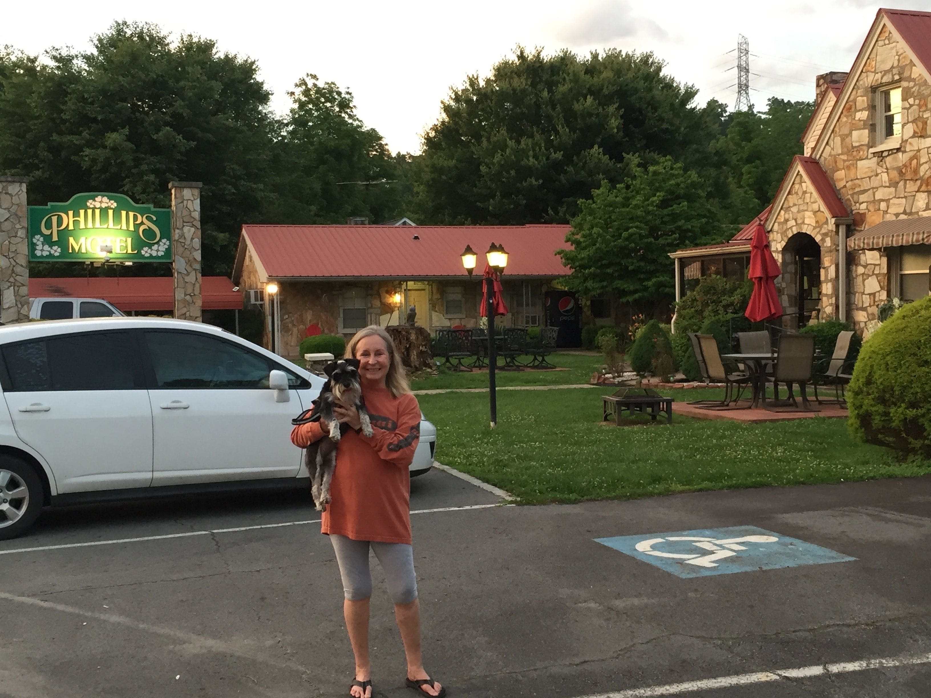 A woman holding a dog in front of a motel with outdoor seating and a stone building.