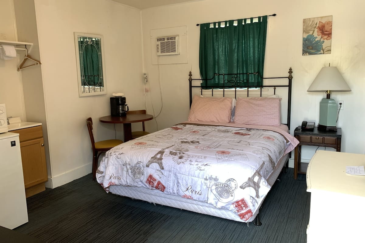 A bedroom with a bed, a desk, a chair, a small table, a lamp, and a radiator.