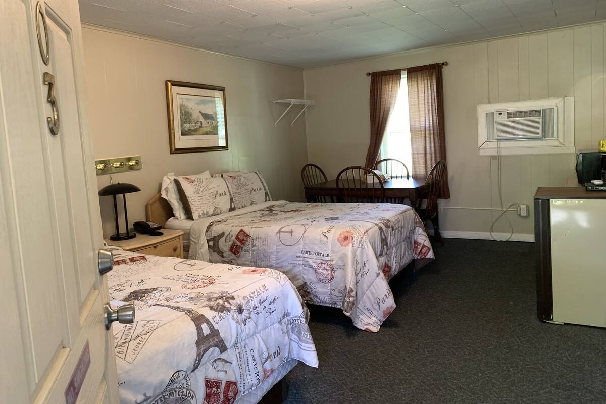 A room with two beds, a desk, a chair, a window with curtains, and a small refrigerator.