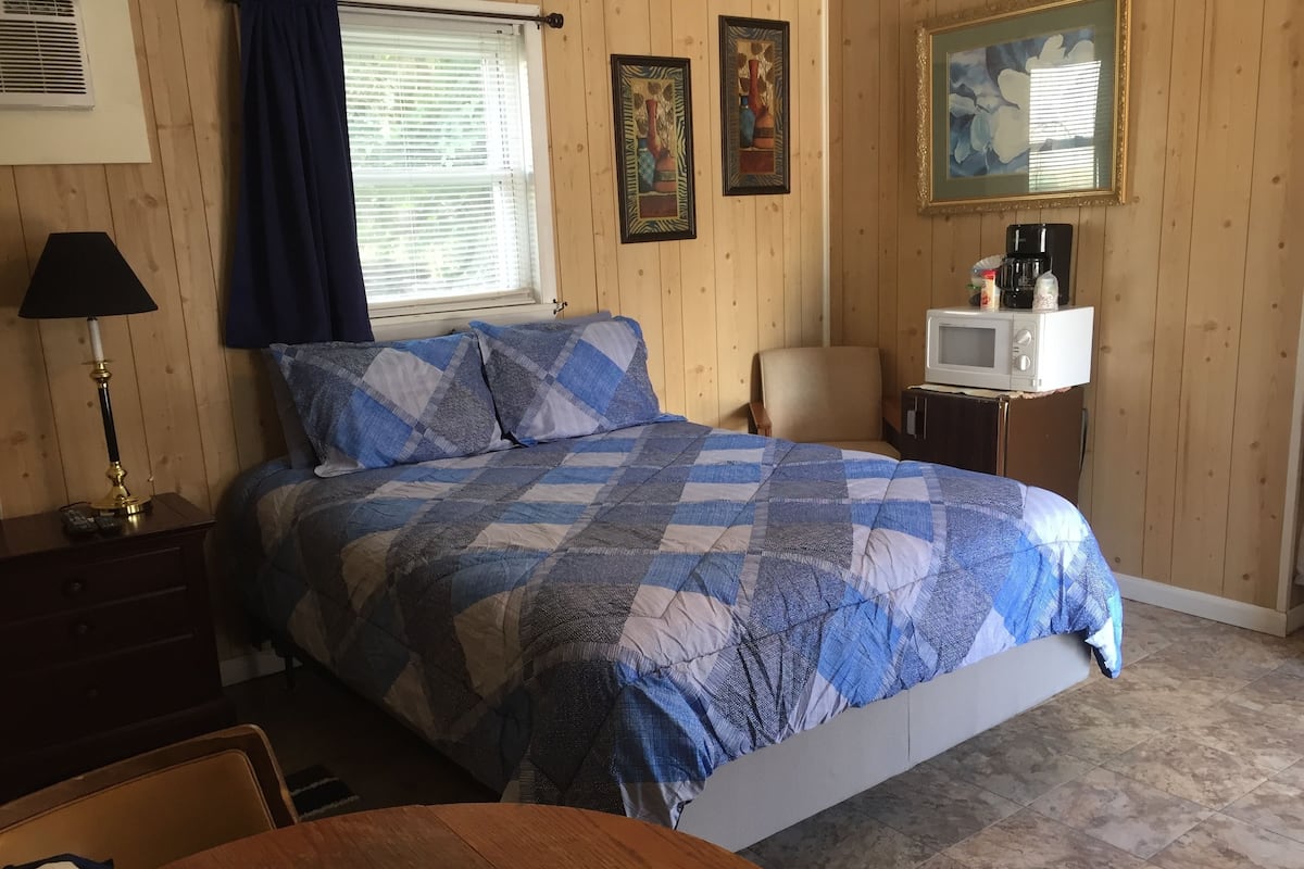 A bedroom with a bed, a desk, a chair, a lamp, a TV, and a coffee maker.