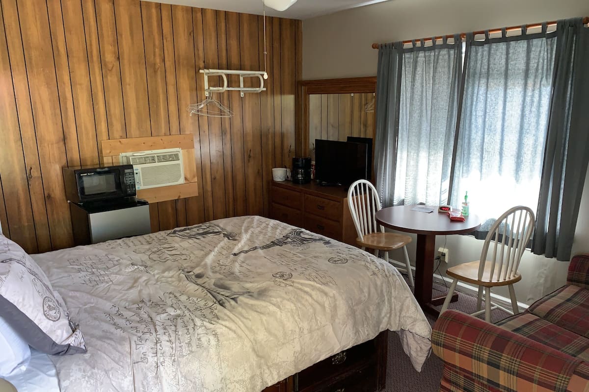 A room with a bed, a wooden wall, a TV, a table with chairs, and a window with curtains.