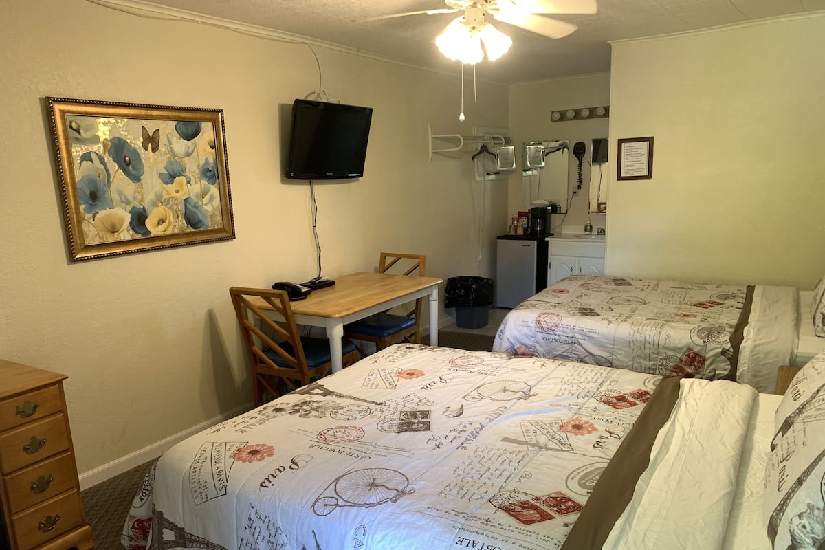 A room with two beds, a desk, a chair, a television, and a ceiling fan.