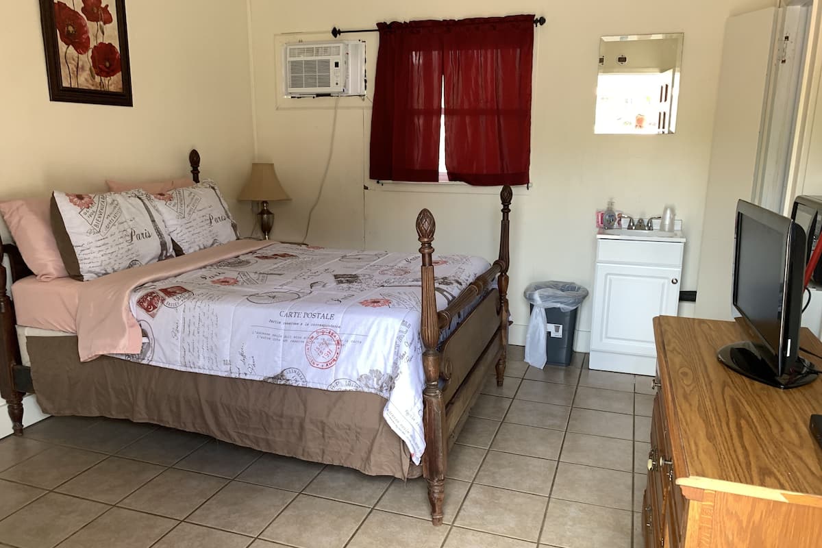 A bedroom with a wooden bed, a bedside table, a TV, a mirror, a window with a red curtain, a wall-mounted air conditioner, and a trash bin.