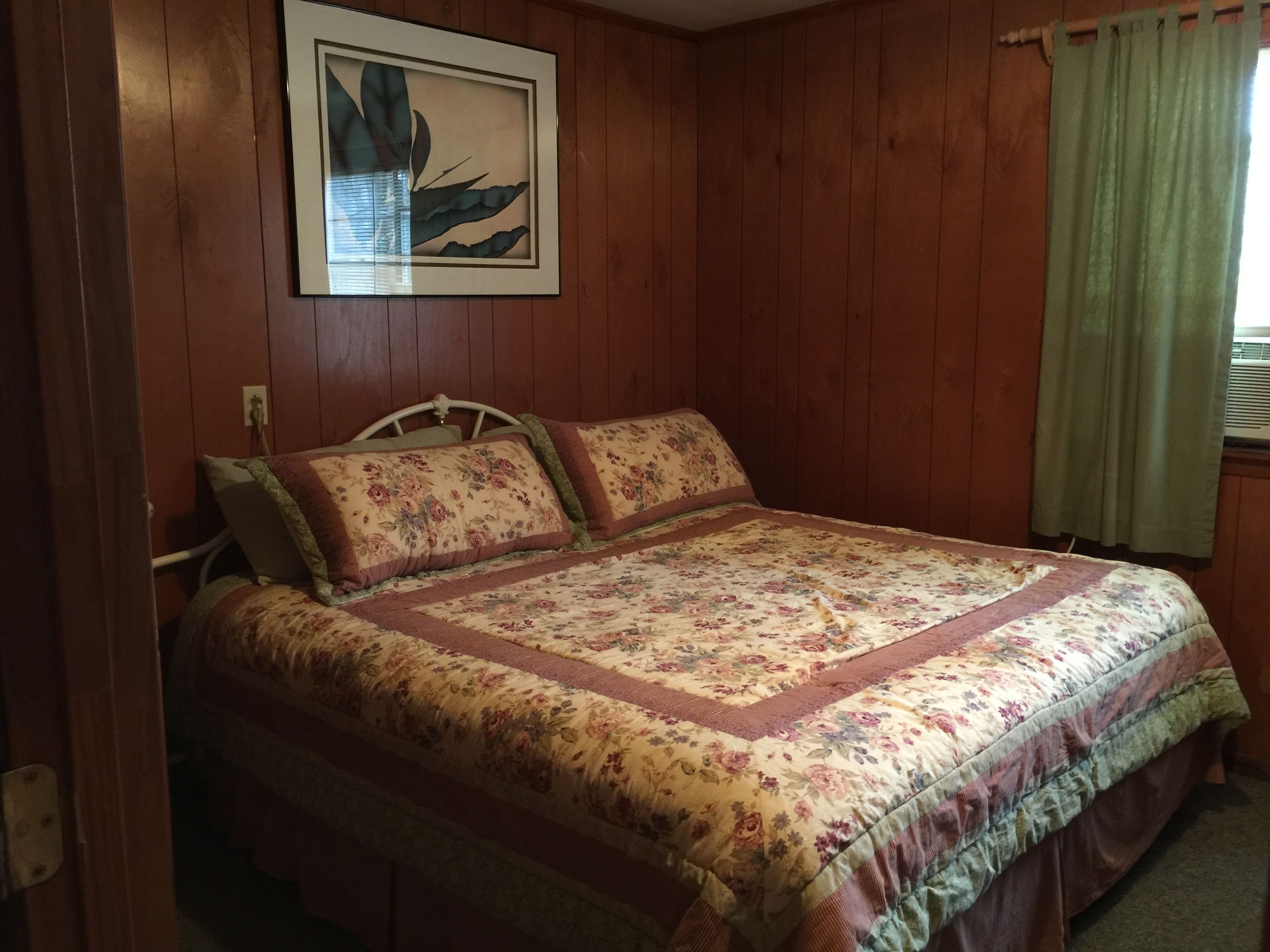A bedroom with a wooden paneled wall, a bed with floral bedding, and a framed artwork above the bed.