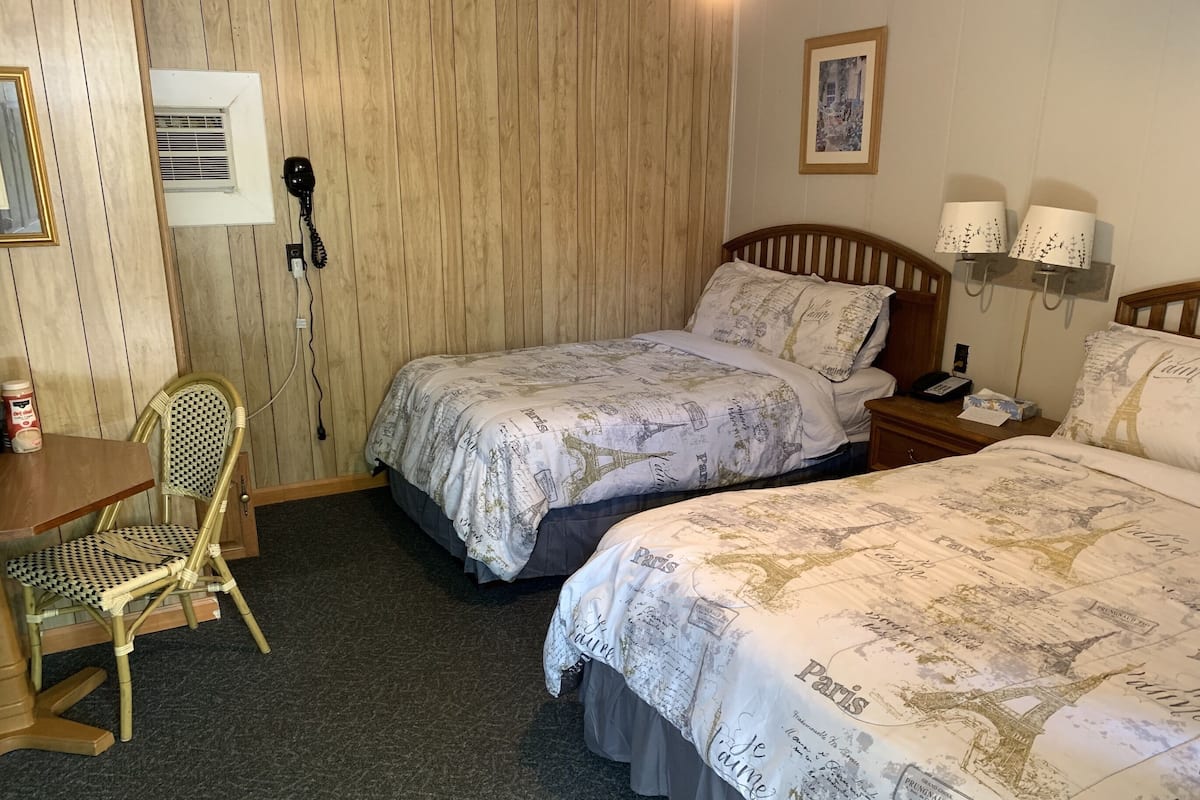 A hotel room with two beds, a wooden chair, a desk, and a wall-mounted phone.