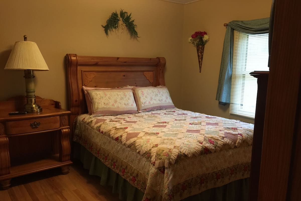 A bedroom with a wooden bed, a nightstand, a lamp, a floral wall hanging, and a window with curtains.