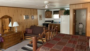 Una habitación acogedora con suelo de madera, una cama con una manta estampada, un aparador de madera, un espejo, una lámpara, un sofá, una mesa de comedor con sillas, un ventilador de techo, una zona de cocina y un baño.