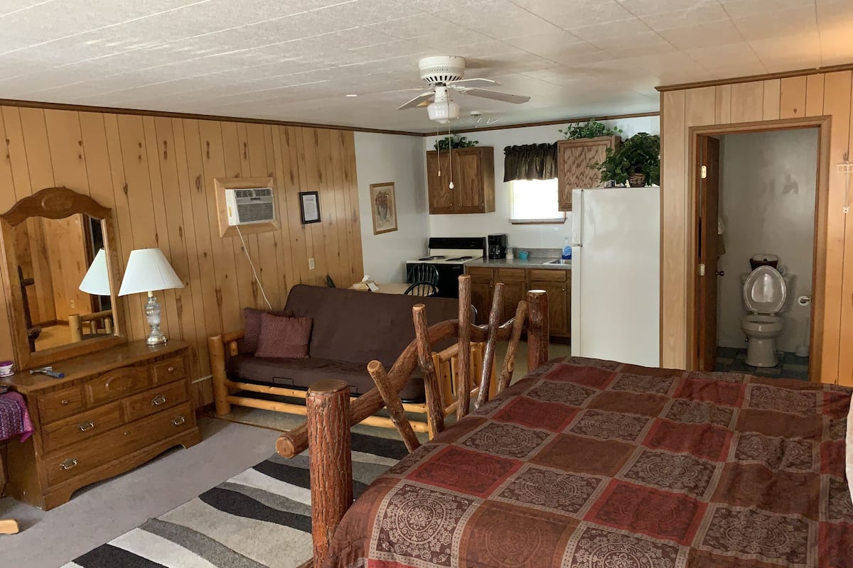 A cozy room with a wooden floor, a bed with a patterned blanket, a wooden dresser, a mirror, a lamp, a sofa, a dining table with chairs, a ceiling fan, a kitchen area, and a bathroom.