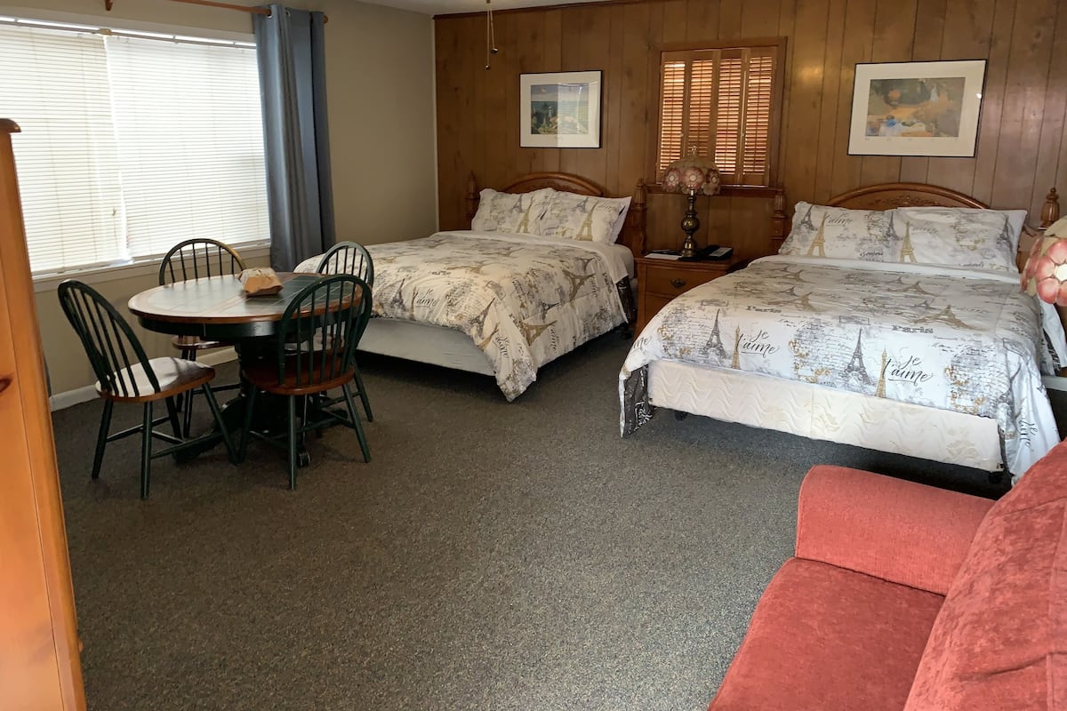 A room with two beds, a dining table with chairs, a ceiling fan, and wooden walls.