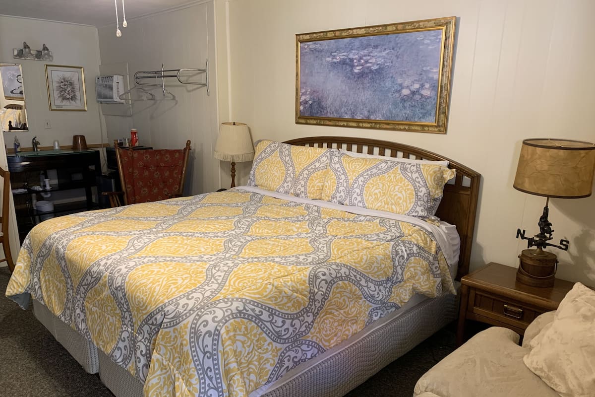 A neatly arranged bedroom with a large bed, a bedside table with a lamp, a small table with a decorative item, and a wall-mounted air conditioner.