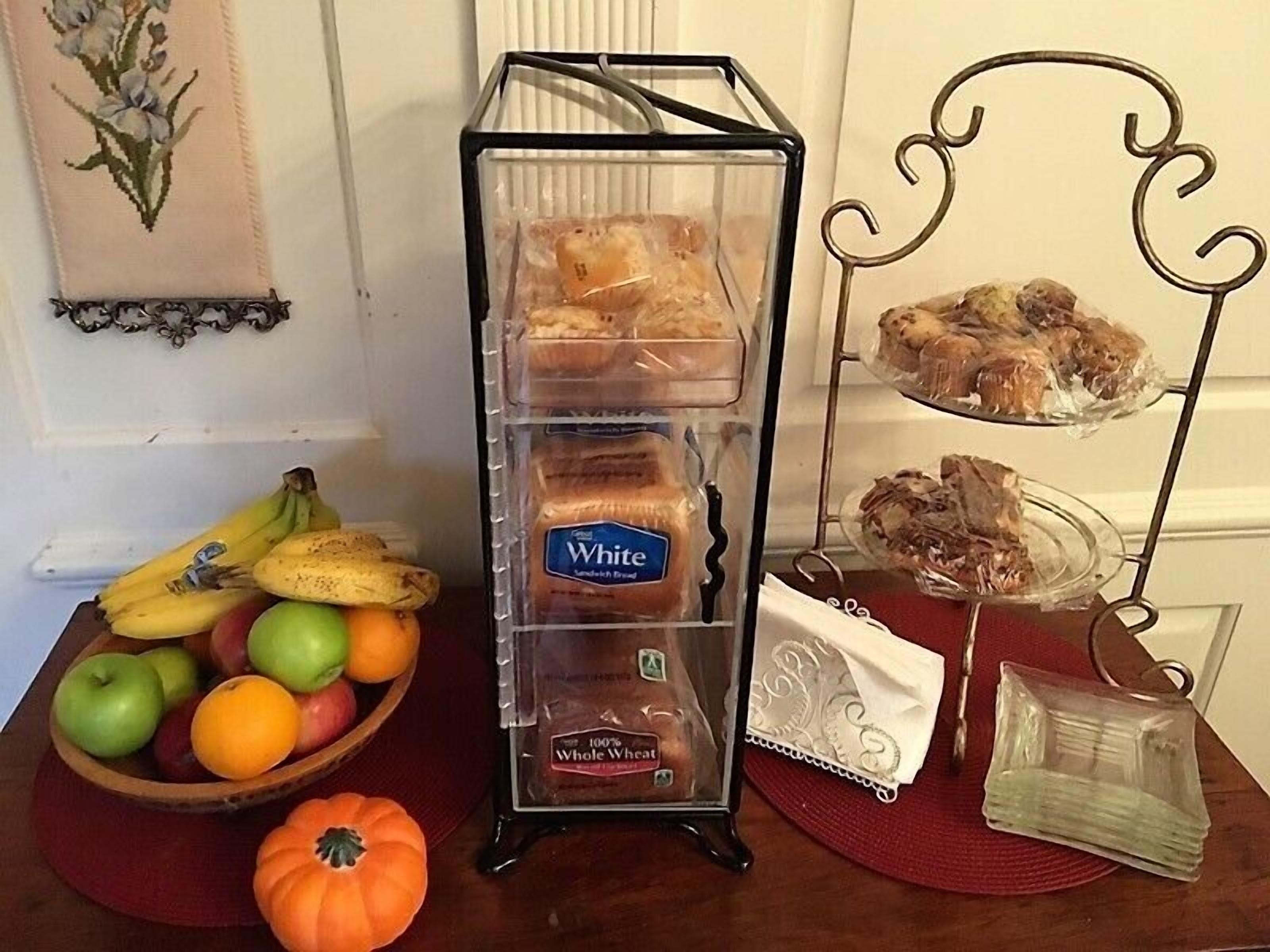 A table with a loaf of White Bread, a bowl of fruit, and a decorative metal stand with pastries.