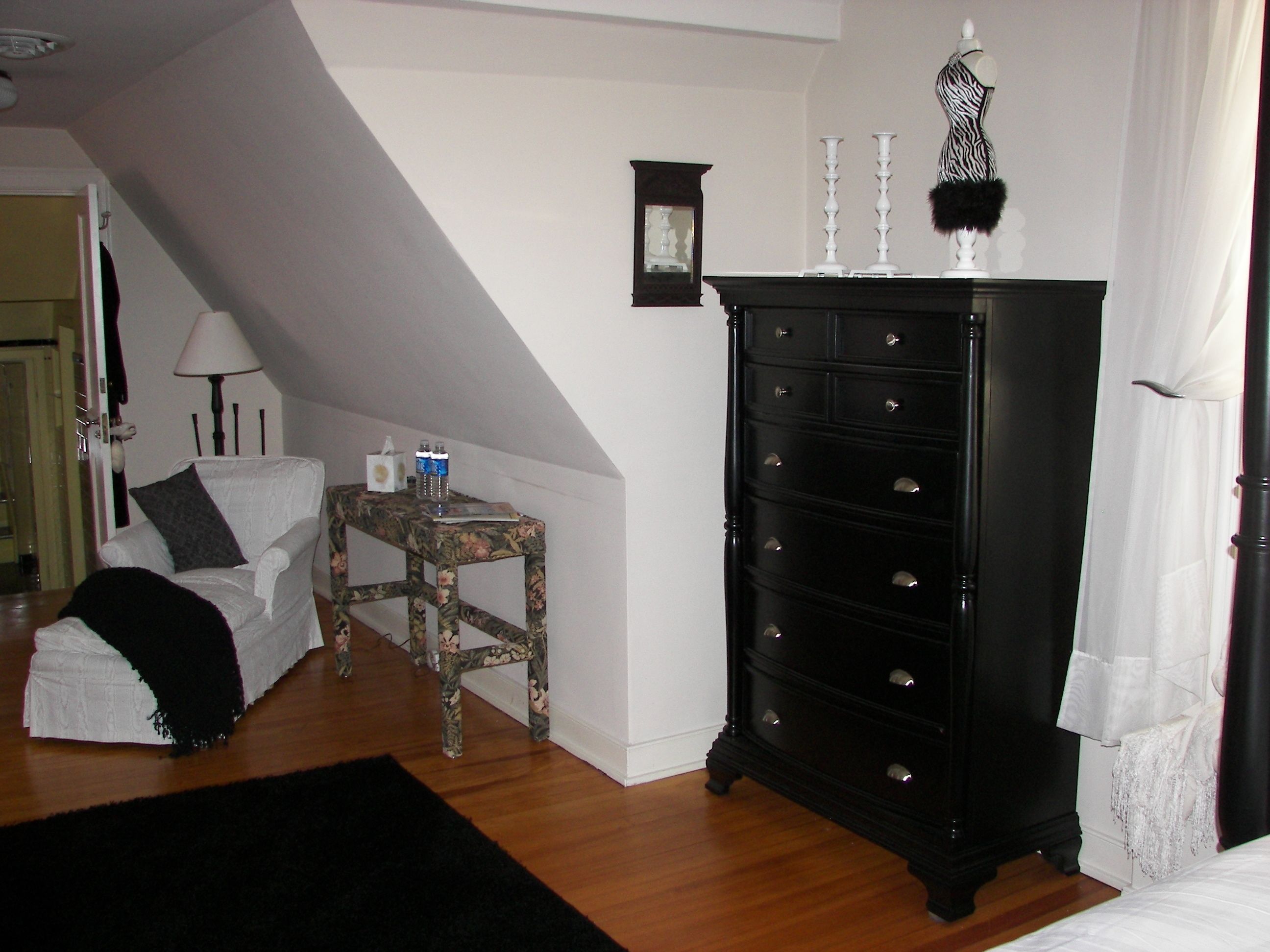 Une chambre à coucher avec un lit blanc, une commode noire, une petite table avec une lampe et une chaise.