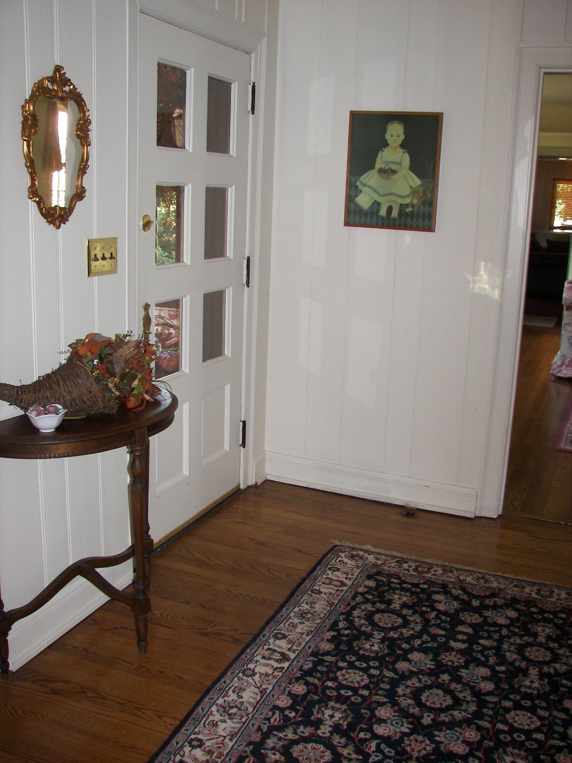 A room with a wooden floor, a white door, a small table with a vase and a bowl, and a patterned rug.