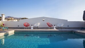 A swimming pool with clear blue water, surrounded by white and red chairs, and a white building with a flat roof in the background. - Aquarius Boutique Hotel (Rethymno)